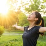 Seorang wanita sedang berjemur di bawah sinar matahari pagi di area taman hijau dengan kedua tangan terbuka, menggambarkan manfaat paparan sinar matahari pagi untuk kesehatan tubuh dan produksi vitamin D alami. (Foto: Ilustrasi/AI)