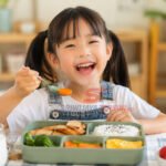 Seorang anak menikmati menu makan siang sehat berisi nasi, ayam, sayuran, telur, dan buah di meja makan dengan suasana ceria. (Foto: dok sriwijayaterkini.co)