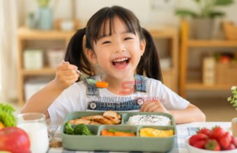 Seorang anak menikmati menu makan siang sehat berisi nasi, ayam, sayuran, telur, dan buah di meja makan dengan suasana ceria. (Foto: dok sriwijayaterkini.co)